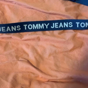 Svart bälte från Tommy Jeans - Snyggt svart bälte från Tommy Jeans med tydlig logga i vitt längs hela bandet. Bältet har detaljer i svart läder och är gjort av textilmaterial. Perfekt accessoar för att lyfta din outfit.