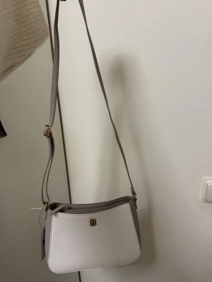 Vit axelväska från Tommy Hilfiger - Snygg vit axelväska från Tommy Hilfiger med justerbart beige axelband och guldfärgad logga framtill. Väskan har en stilren, avlång form och är tillverkad i ett slätt syntetmaterial. Perfekt för dig som vill ha en clean och modern look.