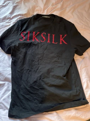 Svart SIKSILK t-shirt med röda stenar - Svart t-shirt från SIKSILK med stora röda bokstäver framtill, dekorerade med glittriga stenar. Klassisk rund hals och korta ärmar. Perfekt för dig som vill sticka ut med en snygg och unik detalj. Oversized i passformen.