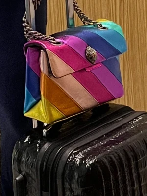 Kurt Geiger väska - Säljer en färgstark axelväska i regnbågsrandigt mönster med metallic-finish. Väskan har kedjerem och coolt spänne framtill. Färgerna går i blått, rosa, gult, orange, lila och grönt. Perfekt statement piece för dig som vill sticka ut. INGA SKAVANKER ELLER NÅGOT- perfekt skick 