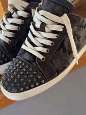 Militär camo loubs - Säljer ett par gråsvarta sneakers från Christian Louboutin med camouflagemönster, vita snören och coola silverfärgade nitar på tån. Skorna har den ikoniska röda sulan och är tillverkade i mocka och skinn. Perfekta för dig som vill sticka ut!