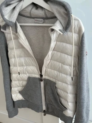 Grå och vit Cardigan från Moncler - Snygg teddyjacka från Moncler i grått och vitt med huva och dragkedja. Jackan har quiltade partier framtill, mjuka muddar och stora fickor. Perfekt för dig som vill ha en mysig och trendig look med exklusiv känsla.