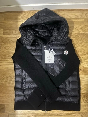 Svart dunväst med huva från Moncler - Snygg svart dunväst från Moncler med huva, ribbstickade svarta ärmar och detaljer. Västen har dragkedja framtill, fickor med dragkedja och Moncler-logga på bröstet. Perfekt för lager-på-lager och streetwear-vibe.
