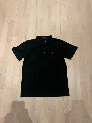 Svart piké från Polo Ralph Lauren - Klassisk svart pikétröja från Polo Ralph Lauren med vit knappslå och den ikoniska röda logotypen broderad på bröstet. Skjortan har korta ärmar och är tillverkad i mjuk bomull som känns skön mot huden.