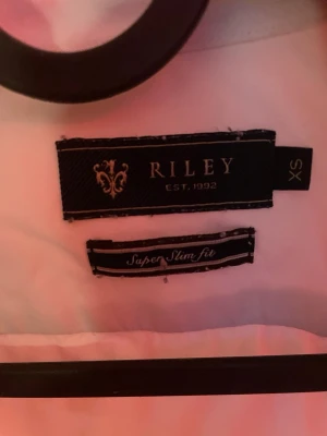Vit skjorta från Riley Super Slim Fit - Klassisk vit skjorta från Riley i super slim fit-modell. Skjortan har lång ärm, krage och knäppning framtill. Perfekt för dig som gillar en tajt och stilren look. Passar till många olika outfits och är ett måste i garderoben.