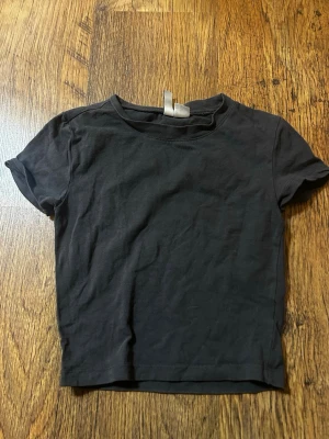 Mörkgrå croppad t-shirt - En enkel mörkgrå croppad t-shirt med rund hals och korta ärmar. Perfekt basplagg att matcha med jeans eller kjol. T-shirten har en klassisk passform och är tillverkad i mjuk bomull för skön komfort.