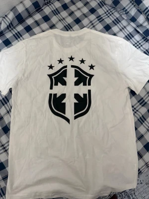 Vit t-shirt med svart grafiskt tryck - Vit t-shirt med ett stort svart grafiskt tryck på ryggen och ett konstnärligt porträtt på framsidan. T-shirten har rund hals och korta ärmar, perfekt för en streetwear-look.