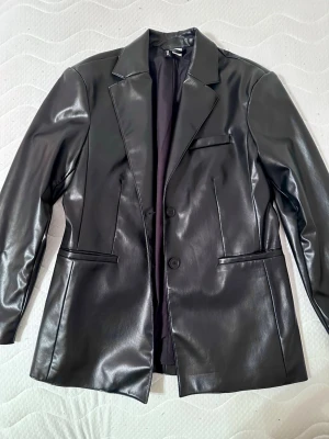 Oversized Leather jacket från Divided H&M - Snygg svart skinnjacka från Divided H&M med klassisk kavajkrage och två knappar framtill. Jackan har raka linjer, fickor på sidorna och en bröstficka för extra stilpoäng. Perfekt för dig som vill ha en edgy look med en clean finish.