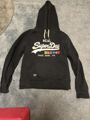 Svart Superdry hoodie med tryck - Svart hoodie från Superdry med stor logga och japansk text i vitt, gult, blått och rött på bröstet. Klassisk känguruficka framtill, ribbade muddar och vit dragsko i huvan. Perfekt för en avslappnad streetwear-look.