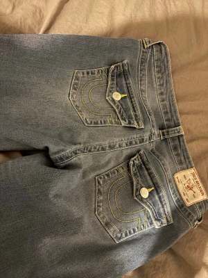 True Religion blå jeans med ficklock - Säljer ett par klassiska blå jeans från True Religion med ikoniska kontrastsömmar och ficklock bak. Jeansen har raka ben och tydliga detaljer på bakfickorna. Perfekta för dig som vill ha en tidlös och snygg denimlook.