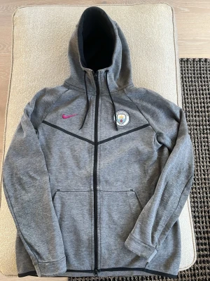 Grå Manchester City hoodie Nike - Snygg grå hoodie från Nike med Manchester City-logga på bröstet och rosa Nike-märke. Dragkedja framtill, två fickor och svart huva med dragsko. Tillverkad i mjukt material, perfekt för dig som gillar sportig stil och vill representera ditt lag. Tyvär syns att den är använd skriv i chatt vid frågor
