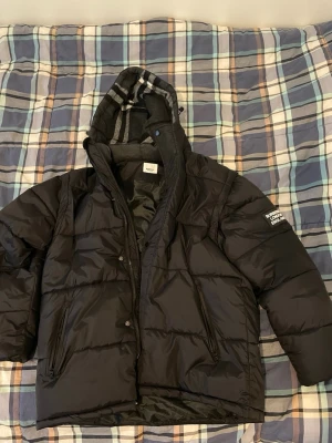 Svart pufferjacka från Burberry - Säljer en svart pufferjacka från Burberry med stor logga på ärmen och klassiskt rutigt foder i huvan. Jackan har dragkedja, tryckknappar och två snedställda fickor framtill. Använt jackan i cirka 1 månad.