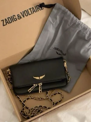 Svart axelväska från Zadig & Voltaire - Snygg svart axelväska från Zadig & Voltaire i skinn med guldfärgade detaljer och kedjerem. Väskan har ett elegant guldemblem med vingar på framsidan och dubbla dragkedjor. Perfekt för dig som vill ha en stilren och trendig accessoar.