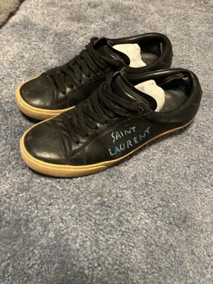 Svarta Saint Laurent sneakers i skinn - Svarta sneakers från Saint Laurent med handskriven logga i blått på sidan. Skorna har snörning, rund tå och en beige platt sula. Tillverkade i skinn med klassisk låg profil som passar till streetwear och casual outfits.