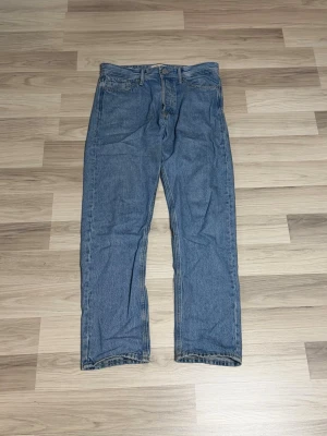 Jack & Jones blåa jeans - Klassiska blå Jack & Jones jeans med rak passform och knappgylf. Jeansen har fem fickor och den ikoniska läderpatchen bak i midjan. Tillverkade i slitstark bomullsdenim som ger en tidlös look.