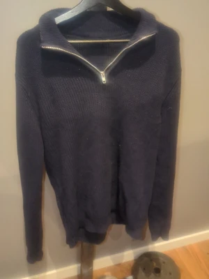 Mörkblå stickad tröja med dragkedja - Mörkblå stickad tröja med hög krage och silverfärgad dragkedja framtill. Tröjan har lång ärm och ribbstickade muddar vid ärmslut och nederkant. Perfekt för lager på lager och en avslappnad stil.