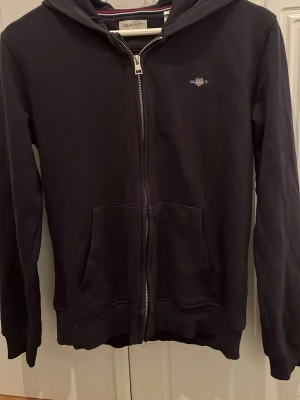 Mörkblå hoodie med dragkedja från Gant, stl 170 - Snygg mörkblå hoodie från Gant med dragkedja framtill och två stora fickor. Klassisk broderad Gant-logga på bröstet och huva med diskreta ränder på insidan. Tillverkad i mjukt bomull/polyester som är skönt att ha på sig.