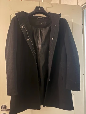 Svart ullkappa med huva  - Stilren svart kappa i ullblandning. Modellen har huva, dold dragkedja och knappar framtill. Perfekt för kyliga dagar och ger en clean look till alla outfits. Längre modell med enkel design och slät yta.