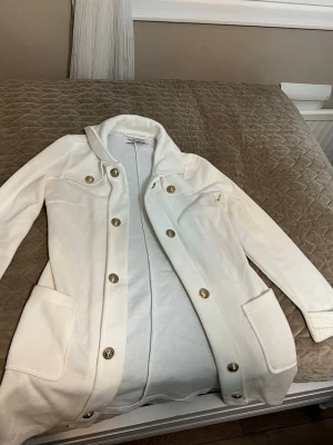 Vit kofta från Chiara Forthi Milano - Stilren vit kofta från Chiara Forthi Milano med guldknappar, två stora fickor framtill och dekorativa bröstfickor. Långärmad modell i mjukt material, perfekt för lager på lager och en clean look.