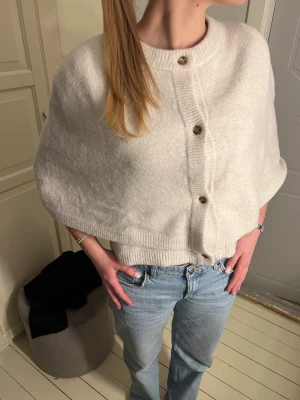 Krämvit cape-kofta med knappar - Snygg krämvit cape-kofta med rund hals och stora bruna knappar framtill.Perfekt att slänga över en t-shirt. Ribbstickade kanter och mjukt, stickat material.Använd endast en gång💓