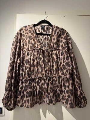 Leopardmönstrad blus från Lindex - Använd 1 gång!!! Trendig blus från Lindex med leopardmönster i beige och bruna toner. Blusen har v-ringning, långa ärmar med resår vid ärmslut och dekorativa knytband framtill.  