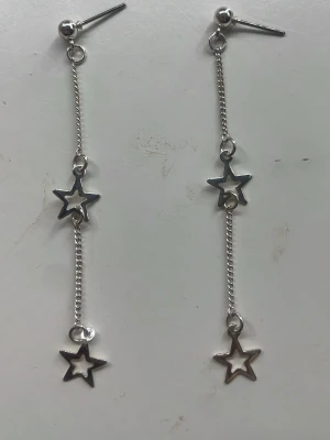 Silvriga stjärnörhängen i kedja - Säljer ett par långa örhängen i silverfärgad metall med två öppna stjärnberlocker på varje örhänge. De hänger i tunna kedjor och har en minimalistisk, trendig look som passar perfekt till en cool outfit.