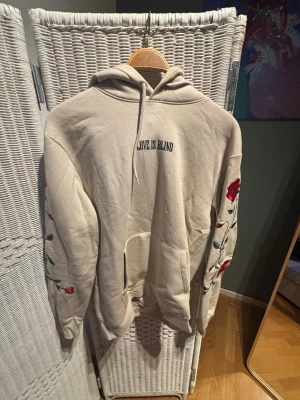 Beige hoodie med rosbrodyr och text från H&M - Säljer en beige hoodie med huva och ficka framtill. På bröstet står det 'LOVE IS BLIND' i svart text och båda ärmarna har broderade röda rosor med gröna blad. Hoodien har en avslappnad passform och är gjord i mjukt bomullsmaterial. Perfekt för en chill och trendig look.