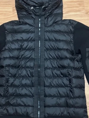 Svart Moncler pufferjacka med stickade ärmar - Säljer en svart pufferjacka från Moncler med stickade ärmar och huva. Jackan har quiltad kropp i blankt material, dragkedja framtill och en liten ficka med dragkedja på ena ärmen. Klassisk Moncler-logga på axeln. STORLEK M