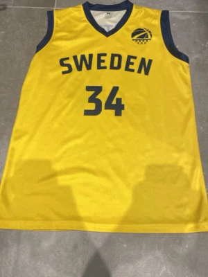 Gul Sweden basketlinne nr 34 S - Säljer ett gult basketlinne från Sweden med marinblå detaljer och nummer 34 på både fram- och baksidan. Namnet Klintman står på ryggen. Tillverkat i 100% polyester, perfekt för dig som gillar basket och vill ha en sportig look.