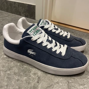 Marinblå mocka sneakers från Lacoste - Snygga marinblå sneakers från Lacoste i mocka med vita detaljer och snörning. Nypris ca 1400kr. Har tyvärr inte kvar kvittot men de köptes från zalando. Pris kan diskuteras! Defekt: Fläck på skosnöret (se sista bild).