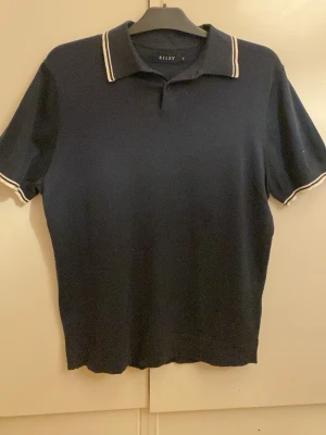 Marinblå polotopp från Riley - Klassisk marinblå polotopp från Riley i storlek M. Toppen har korta ärmar, krage med vita ränder och knäppning framtill. Tillverkad i ett mjukt stickat material som känns skönt mot huden. Perfekt för en stilren och enkel look.