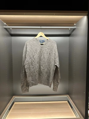 Grå stickad tröja från Polo Ralph Lauren - Klassisk grå stickad tröja från Polo Ralph Lauren i 100% lammull. Tröjan har v-ringning, långa ärmar och den ikoniska broderade loggan i vinrött på bröstet. Perfekt för lager på lager under höst och vinter. (Liten defekt på bröstet)                                                                             Längd:62cm.     Bredd:44cm