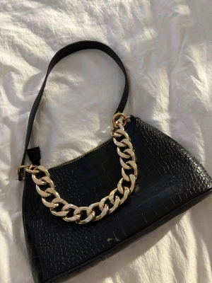 Svart axelväska med kedjedetalj - Säljer en svart axelväska i croco-mönstrad syntet med en chunky guldfärgad kedja framtill. Väskan har dragkedja upptill och en enkel axelrem. Perfekt storlek för det viktigaste och ger en cool vibe till din outfit.