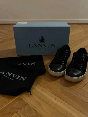 Lanvins skor svarta - Säljer ett par svarta sneakers från Lanvin med vit platt sula och klassisk snörning. Skorna har en stilren och tidlös design, perfekta till jeans eller chinos. Kommer med originalkartong och dustbags. Pris kan diskuteras 💸
