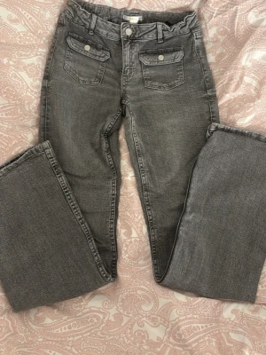 Gråa bootcut jeans med fickor - Säljer ett par gråa bootcut jeans med två stora fickor framtill som har knappar. Jeansen har bälteshällor och klassisk knappgylf. 