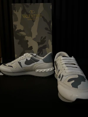 Valentino Rockrunner sneakers camo - Snygga Valentino Rockrunner sneakers i vitt och grått med camouflagemönster. Skorna har coola nitar på hälen, vita snören och en mix av skinn och textil. Perfekt för dig som vill sticka ut med exklusiva detaljer och streetstyle-vibe.