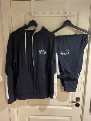 Hugo boss Treksut - Säljer ett par marinblå mjukisbyxor från BOSS med vita detaljer längs sidorna och logga tryckt på benet. Byxorna har resår i midjan med snörning och muddar vid bensluten. Perfekta för chill eller träning.