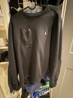 Svart sweatshirt från Polo Ralph Lauren - Svart sweatshirt från Polo Ralph Lauren med meshficka och dragkedja på bröstet samt broderad logga. Tröjan har långa ärmar med detaljer i annat tyg och ribbade muddar. Perfekt för dig som gillar stilrena och sportiga plagg.