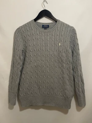 Ralph Lauren kabelstickad - Ljusgrå Polo Ralph Lauren-tröja i kabelstickad bomull, storlek L(14-16), mycket bra skick utan hål eller fläckar. Klassisk rund hals med broderad polospelare och ribbade kanter, unisex. Vid frågor är det bara att höra av dig😄