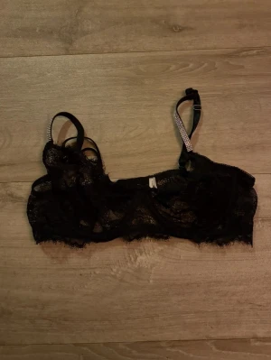 Svart spetsbralette med strassband - Säljer en svart bralette i spets med tunna axelband dekorerade med strassdetaljer. Modellen har genombruten spets och en feminin look med fina kanter. Perfekt för dig som gillar detaljer och vill ha något extra.