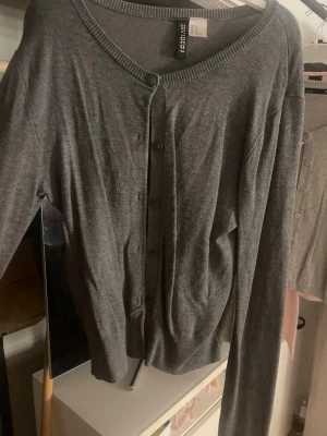 Grå kofta från Divided - Säljer en enkel och stilren grå kofta från Divided. Koftan har långa ärmar, rundad halsringning och knappar framtill. Perfekt att slänga över en t-shirt eller linne för en chill look. Materialet är mjukt och tunt, troligen bomull eller polyester.