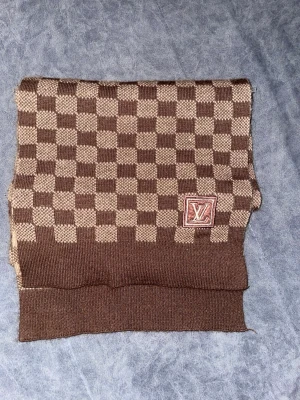 Lv halsduk - (Pris kan diskuteras) Säljer en brun och beige rutig halsduk från Louis Vuitton med klassiskt schackrutigt mönster och LV-logga framtill. Perfekt för dig som vill ha en ikonisk och stilren look.