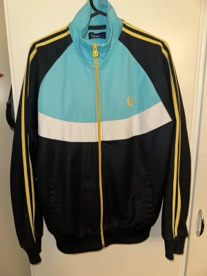 Fred Perry retro vindjacka M - Säljer en retroinspirerad vindjacka från Fred Perry i storlek M. Jackan har svart botten med ljusblå och vit panel, gula detaljer och dragkedja samt klassiska ränder på ärmarna. Hög krage och ikonisk Fred Perry-logga på bröstet. Perfekt för dig som gillar sportig stil.