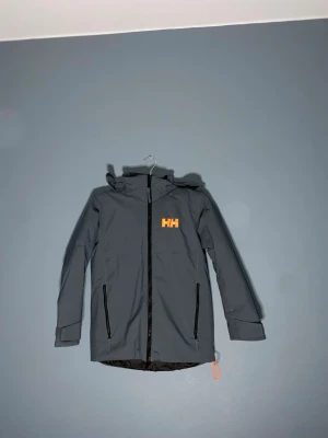 Mörkgrå vindjacka från Helly Hansen - Säljer en mörkgrå vindjacka från Helly Hansen med orange HH-logga på bröstet och huvan. Jackan har huva, två dragkedjeförsedda fickor och justerbar nederkant med orange snodd. Perfekt för blåsiga dagar och outdoor.