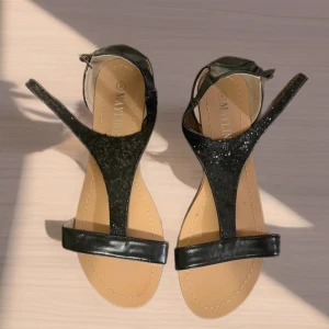 Svarta glittriga sandaler från Mayline - Säljer ett par svarta sandaler från Mayline med glittriga detaljer på remmarna. Skorna har öppen tå, platt sula och justerbart spänne vid ankeln. Perfekta för sommarens alla tillfällen och ger en elegant touch till din outfit.