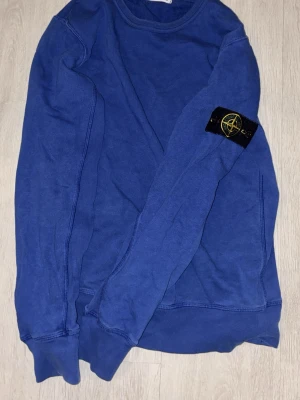 Blå  tröja från Stone Island - Säljer en blå stickad tröja från Stone Island med den ikoniska kompassen på ärmen. Tröjan har rund halsringning och långa ärmar. Perfekt för dig som vill ha en clean och stilren look med streetwear-vibe.