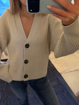 Beige ribbad kofta från H&M - Säljer en beige ribbad kofta från H&M i storlek M. Koftan har stora, snygga knappar framtill och en djup v-ringning. Den är stickad med breda muddar vid ärmslut och nederkant, perfekt för lager på lager under höst och vinter.