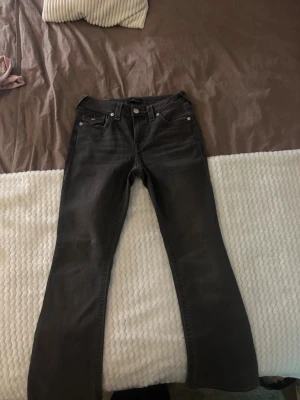 True Religion mörkgråa bootcut jeans - Mörkgråa jeans från True Religion, modell Becca  Rise Bootcut. Snygga kontrastsömmar och ikoniska fickdetaljer med silverknappar bak. Klassisk femficksdesign och bootcut-ben för en chill vibe.Tycker de sitter som en lite förhöjd low waist. Uppsydda lite men sitter perfekt på mig som är 166cm. Använda 3ggr