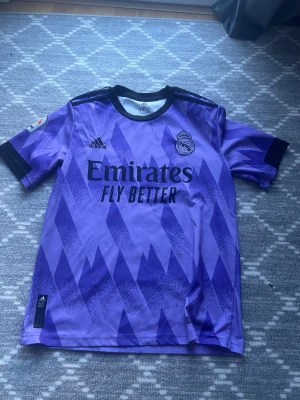 Fotbollströja  - Säljer en lila Real Madrid matchtröja från Adidas med Benzema och nummer 9 på ryggen. Tröjan har svart krage, svarta detaljer på ärmarna och ett grafiskt mönster i olika lila nyanser. Tillverkad i lätt och ventilerande material, perfekt för fotboll eller som streetwear.
