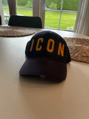 Dsquared - Snygg marinblå keps från Dsquared2 med stora gula bokstäver 'ICON' framtill och 'DSQUARED2' broderat baktill. Använd fåtal gånger, skick 9/10. Tillverkad i bomull för en bekväm känsla och cool look. Nypris ca 1300.